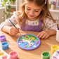 Young girl painting a colourful unicorn craft plaque with a paintbrush at a wooden table.