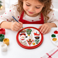 Young girl painting a round Christmas craft plaque featuring Santa Claus, a snowman and Christmas tree.