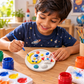 Child painting a round space-themed DIY craft with astronauts, rocket and moon using colourful paints and a brush.